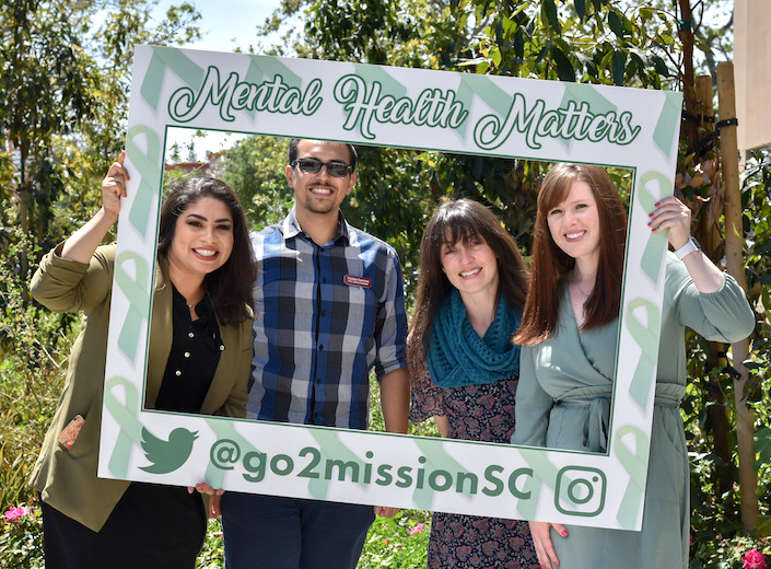 Four people lean into a prop photo frame that reads "Mental health matter!" for a photo outside on campus.