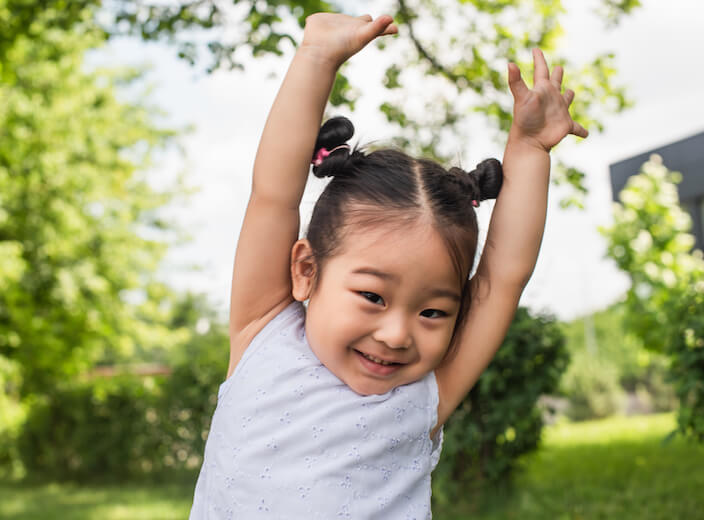 Asian toddler with her hands in the air. She wears her hair in short pigtails and smiles.