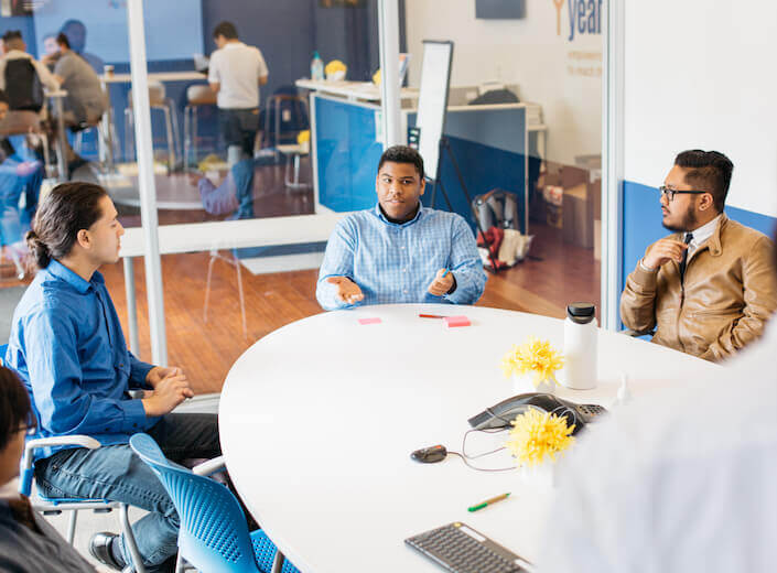 Young adult coworkers work together in a modern office. Three of them sit together at a table and wear business casual clothes.