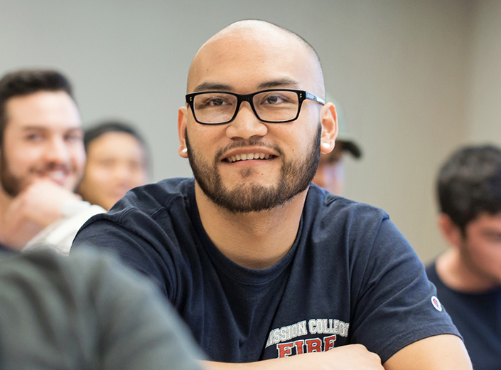 EMT training. A young man with a shaved head and black glasses smiles at a presentation in a classroom.