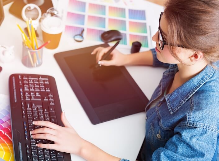 A young woman with brown hair pulled back in a ponytail works at a white desk on a laptop. Creative supplies surround her.