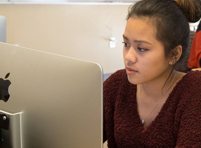 Adobe Illustrator. A young woman with long black hair pulled back in a ponytail is working at a desktop computer.