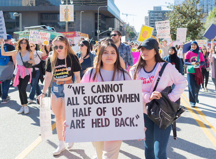 A young woman in Los Angeles holds a sign up that reads "We cannot all succeed when half of us are held back." Various types of women are seen behind her, holding signs and walking.