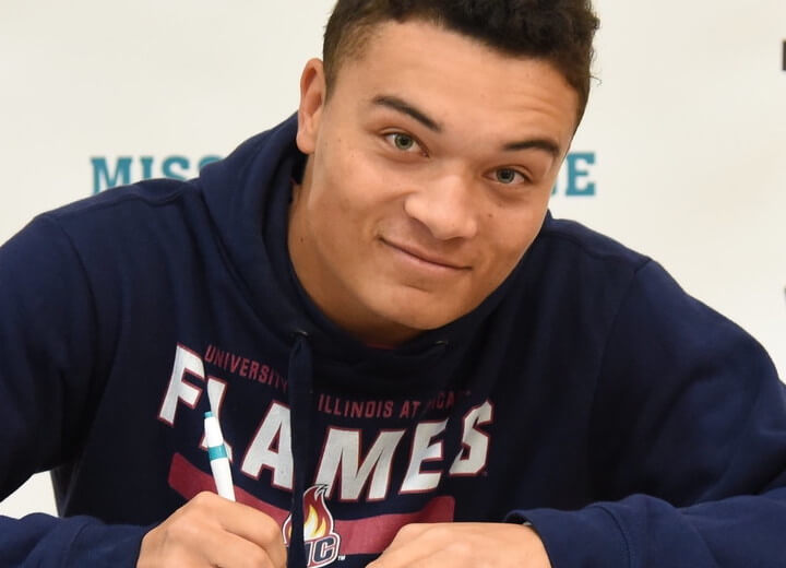 Young man with light brown skin, short curly hair, and blue eyes wears an athletic sweatshirt holds a pen and signs a piece of paper in a ceremony about his transfer to a four-year college.