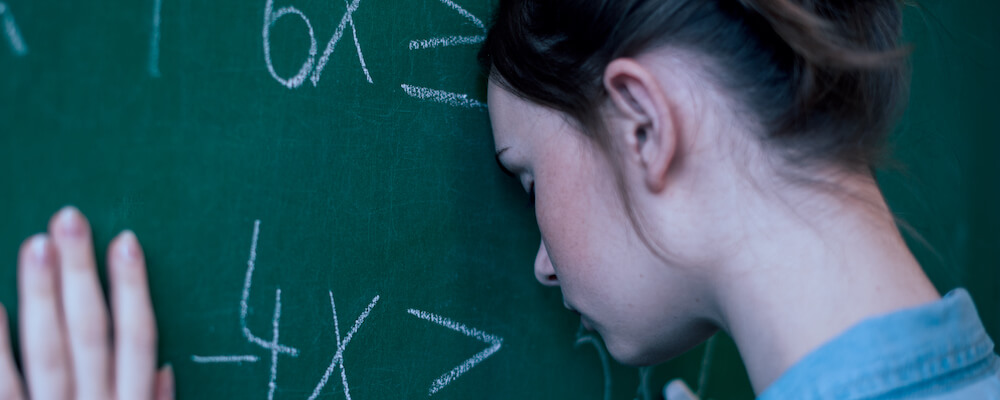 A young woman with light skin and brown hair pulled back in a bun leans against a blackboard (forehead is touching it) that has equations written on it.