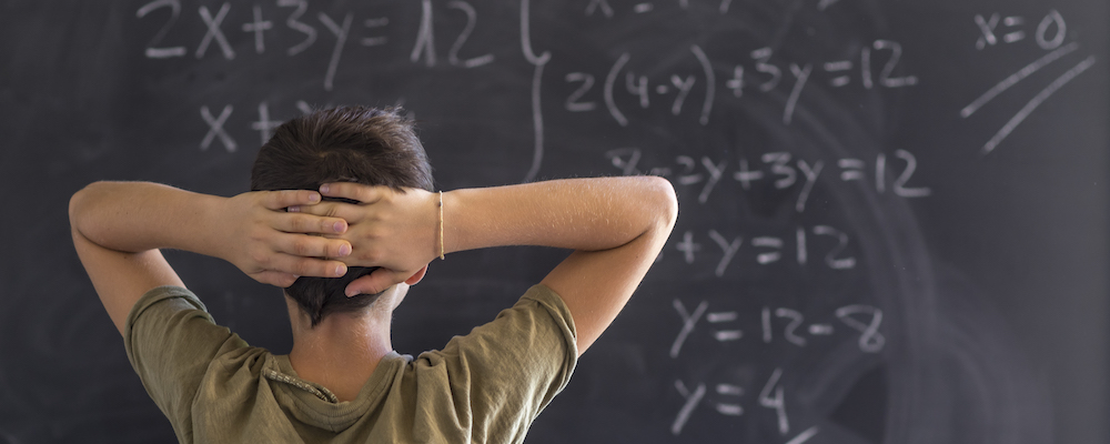 A male student with short brown hair crosses his arms behind his head and contemplates a math problem at the blackboard in his classroom.