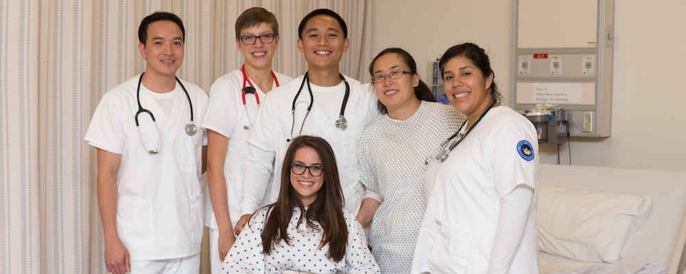 Student nurses pose in their white scrubs in the simulation lab. A patient in a white hospital gown sits in a wheelchair in front of the group.