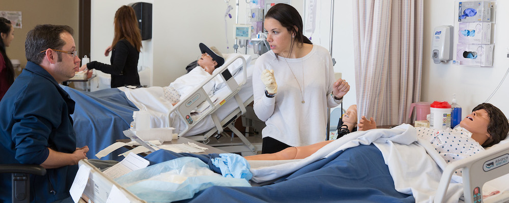 Nursing faculty works with student who is administering care in scrubs to a dummy patient in a hospital bed.