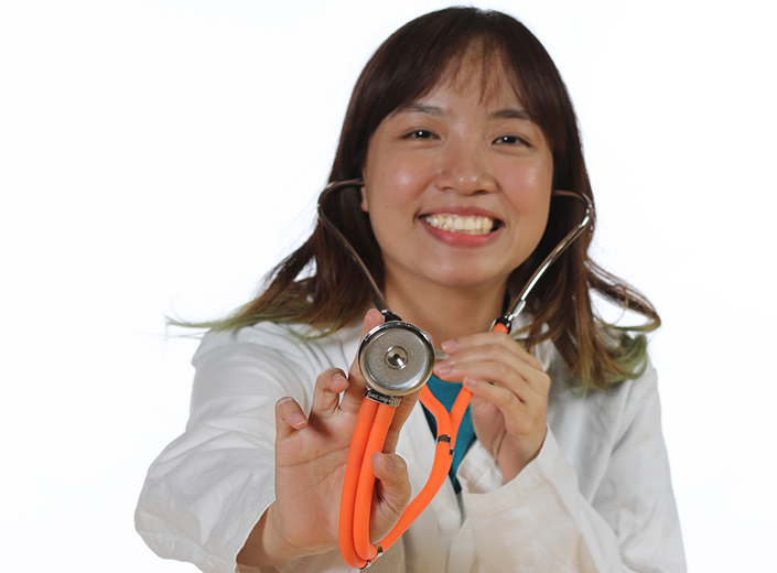 Student nurse in white coat holds stethoscope up to camera. She is of Asian descent and has straight black hair to her shoulders.