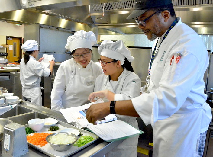 Instructor chef works with two female students in the kitchen.