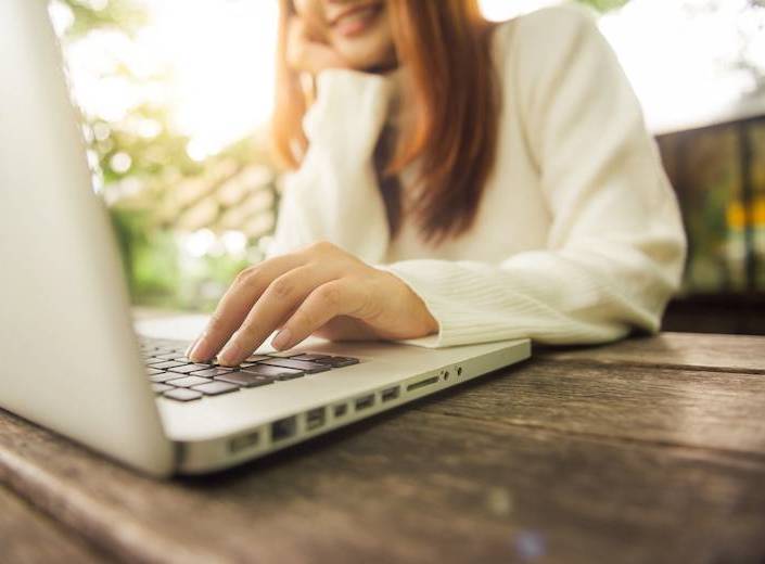 Girl with long-sleeved shirt and straight brown hair works at a laptop. Her face is out of view.