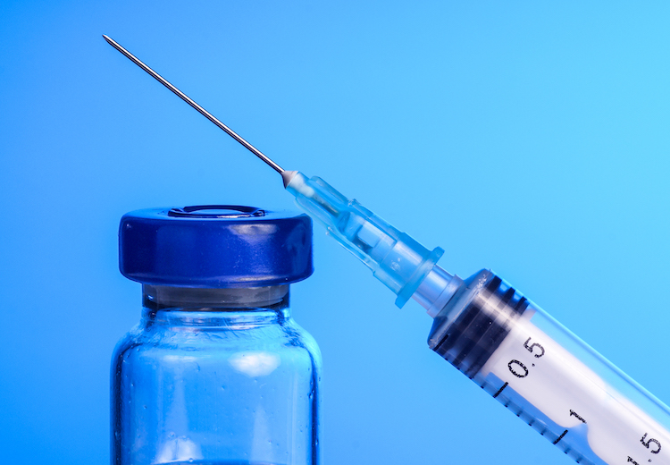A needle and small clear glass bottle of vaccine are pictured against a deep blue background.
