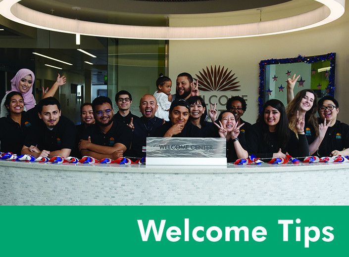 Welcome is written on teal strip at the bottom of an image of a group of several students posted behind the counter of the desk of the Welcome Center.
