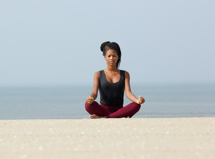 An female African-American college student with long braids tied on the top of her head is sitting with her legs crossed and hands resting on each knee. She is on the beach.