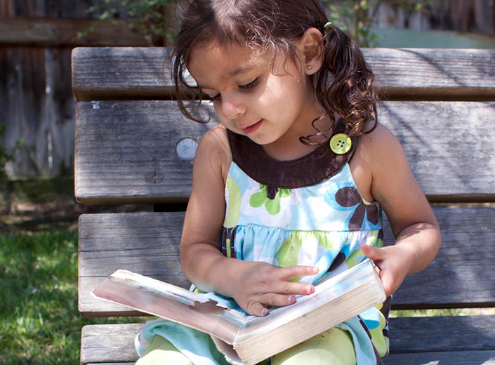Young girl with curly brown hair reads a book on a park bench.