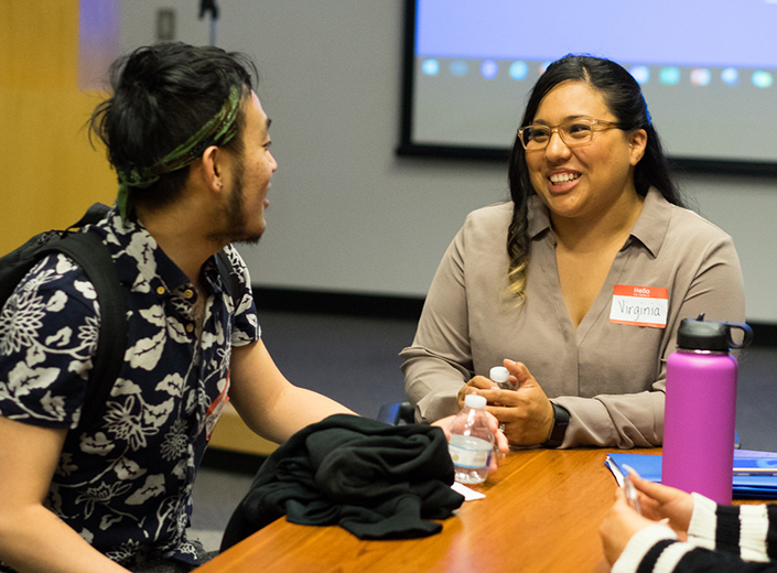 Latina with name tag, long black hair, glasses smiles at male student. They sit at a table in a classroom.