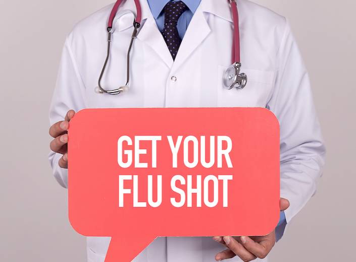 A doctor in a white coat is holding a bulletin board that reads "Flu Shots".