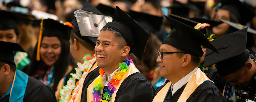 Two young men wear commencement cap and gowns and sit in a crowd of other graduates in an auditorium.