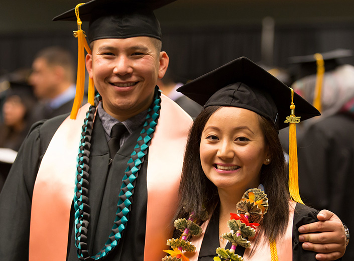 A male and female student pose together at graduation in their commencement cap and gowns. They have various sashes and ribbons on denoting their involvement with various organizations on campus.