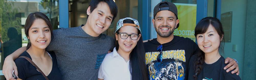 Four students in tshirts pose with their arms around each other in front of a building on campus.