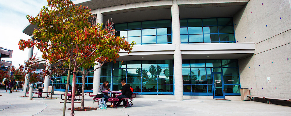 Campus Center from the outside, a modern looking building with lots of tinted glass windows. Two students sit outside at a table.