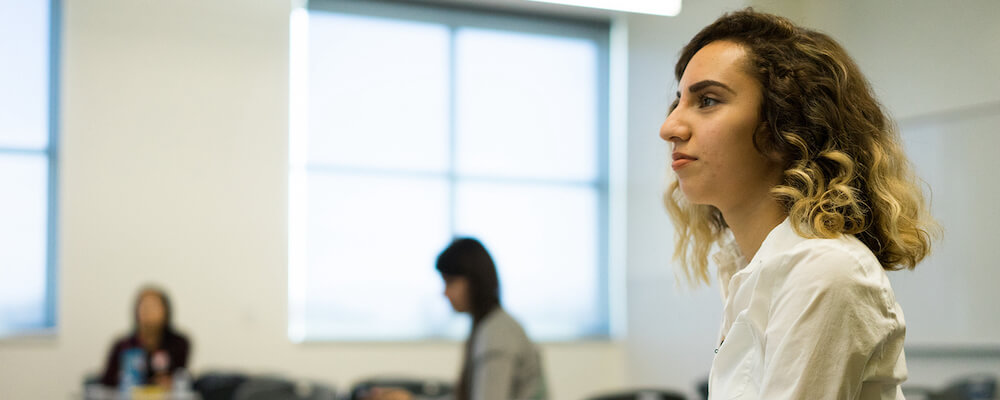 Young woman with curly light brown hair and olive skin sits in a classroom. She is pictured in profile, and she looks toward the front of the room.