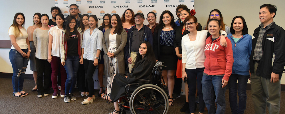 Group of EOPS students are commencement in their casual clothes. They stand in front of an EOPS wall banner.