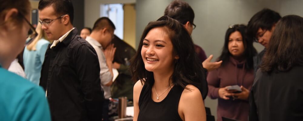 Young woman with shoulder-length black hair mingles at a campus event. She is laughing andwears a sleeveless black shirt and name tag.
