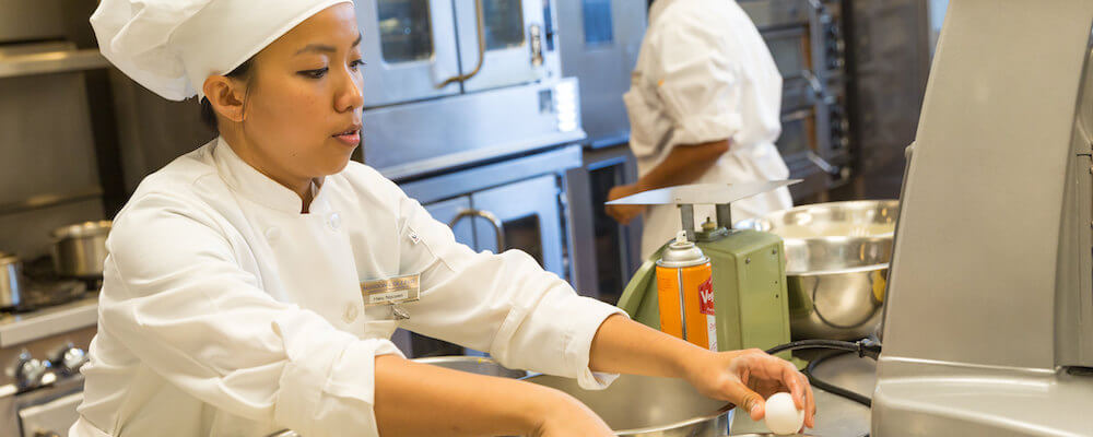 Female student of Asian-American descent wears a white chef uniform and prepares food using several metal bowls in a kitchen.