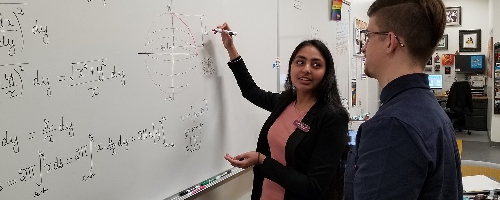 A young woman with long dark hair in a blazer and name tag tutors a male student in math at the marker board in a classroom.