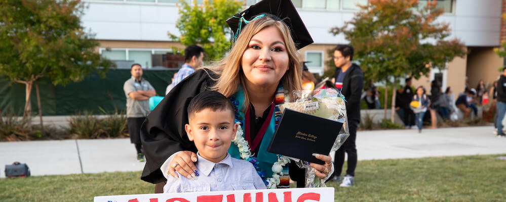 Latinx mom and son at commencement. The mom is youngish and wears a cap and gown. She has light brown hair. Her son holds a sign that says "Congrats Mom!"