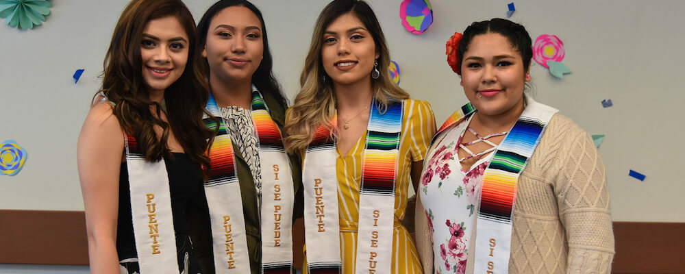 Cuatro mujeres jóvenes se paran juntas en un evento de graduación. Llevan fajas que dicen "Puente". Las fajas tienen hilos multicolores en verde, amarillo, naranja, rojo, azul, blanco y negro.