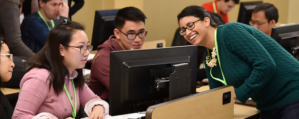 Students work in a computer lab. A female instructor with dark hair in a ponytail leans around the desk to demonstrate something to a female student in glasses.