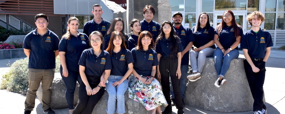 Group of students in jeans and sweatshirts pose in front of the Welcome Center on campus.