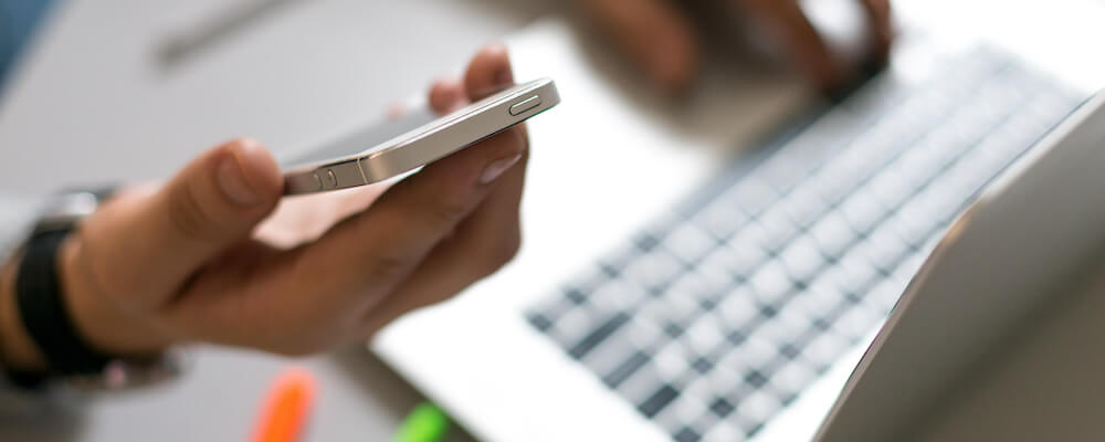 A pair of hands with light skin, possibly female, hold a smartphone and work at an Apple laptop. A few colorful pens are scattered nearby.