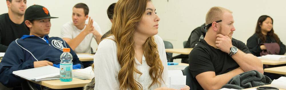 Students in a classroom. The young woman in the center of the picture has long light brown hair and wears a white shirt. She is LatinX.