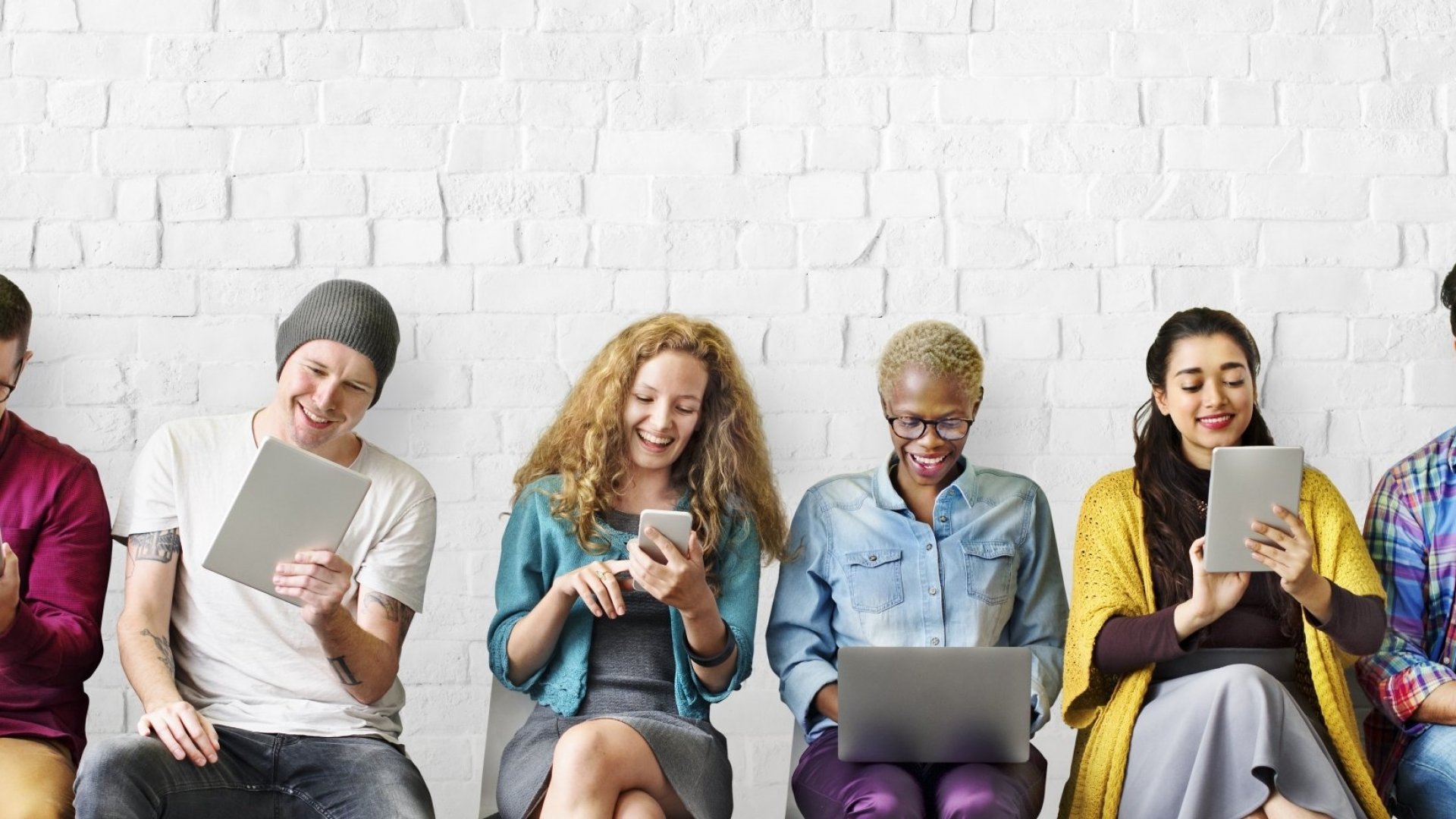 Young adults sit in chairs and look at phones, tablets, computer.s They look happy.