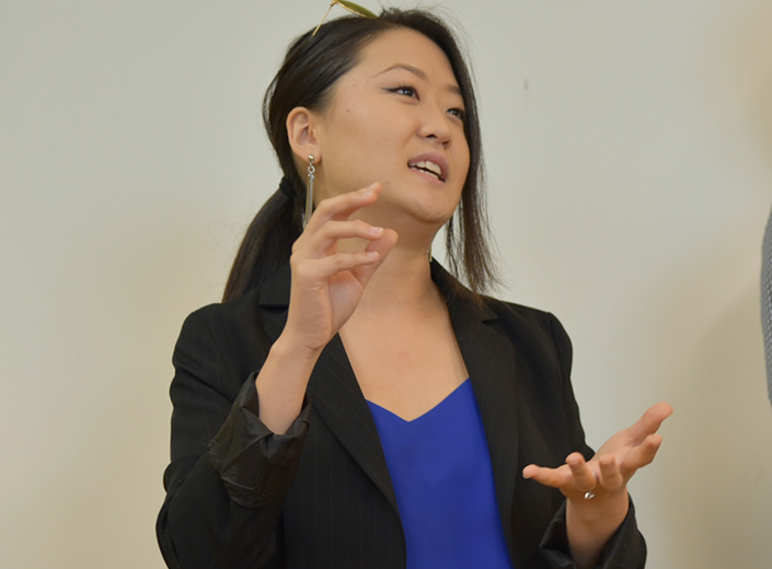 Female student of Asian descent delivers a speech. She wears a black blazer and blue shirt.